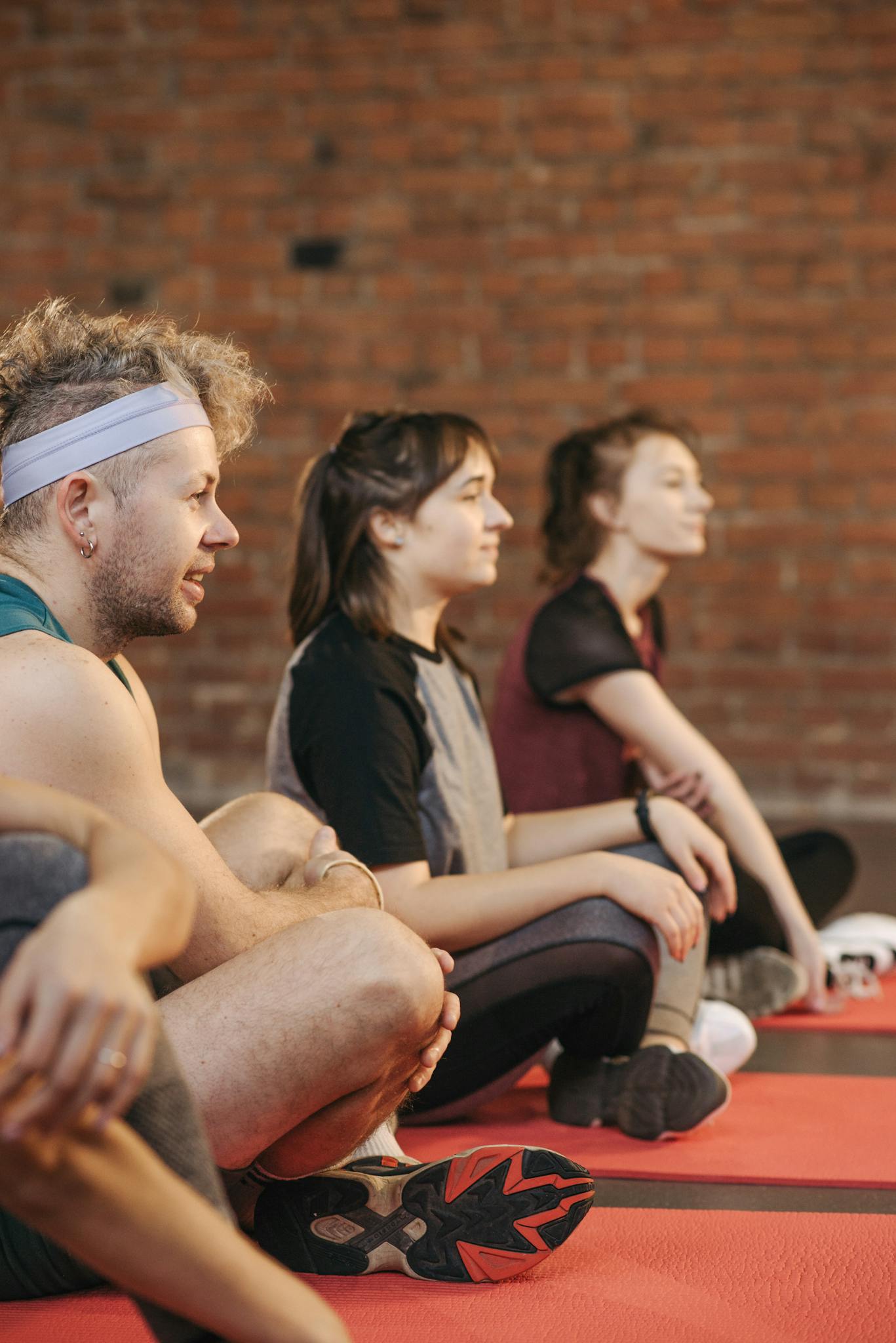 Three adults in a gym setting during a yoga session, seated on red mats.