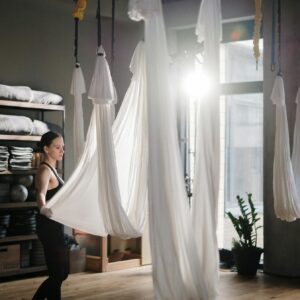 A woman participates in an aerial yoga session in a well-lit studio with hanging silks.