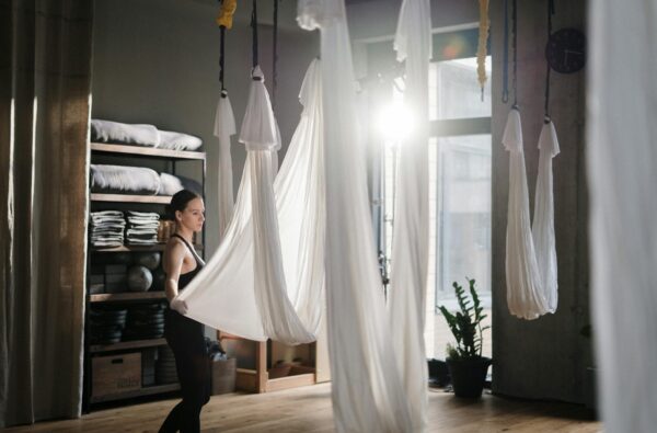A woman participates in an aerial yoga session in a well-lit studio with hanging silks.
